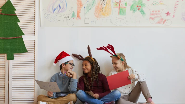 Three children sit on the floor wearing festive hats and reindeer antlers, smiling and holding papers. Behind them is a wall decorated with colorful holiday drawings, capturing the joyful spirit of homeschooling through the holidays.
