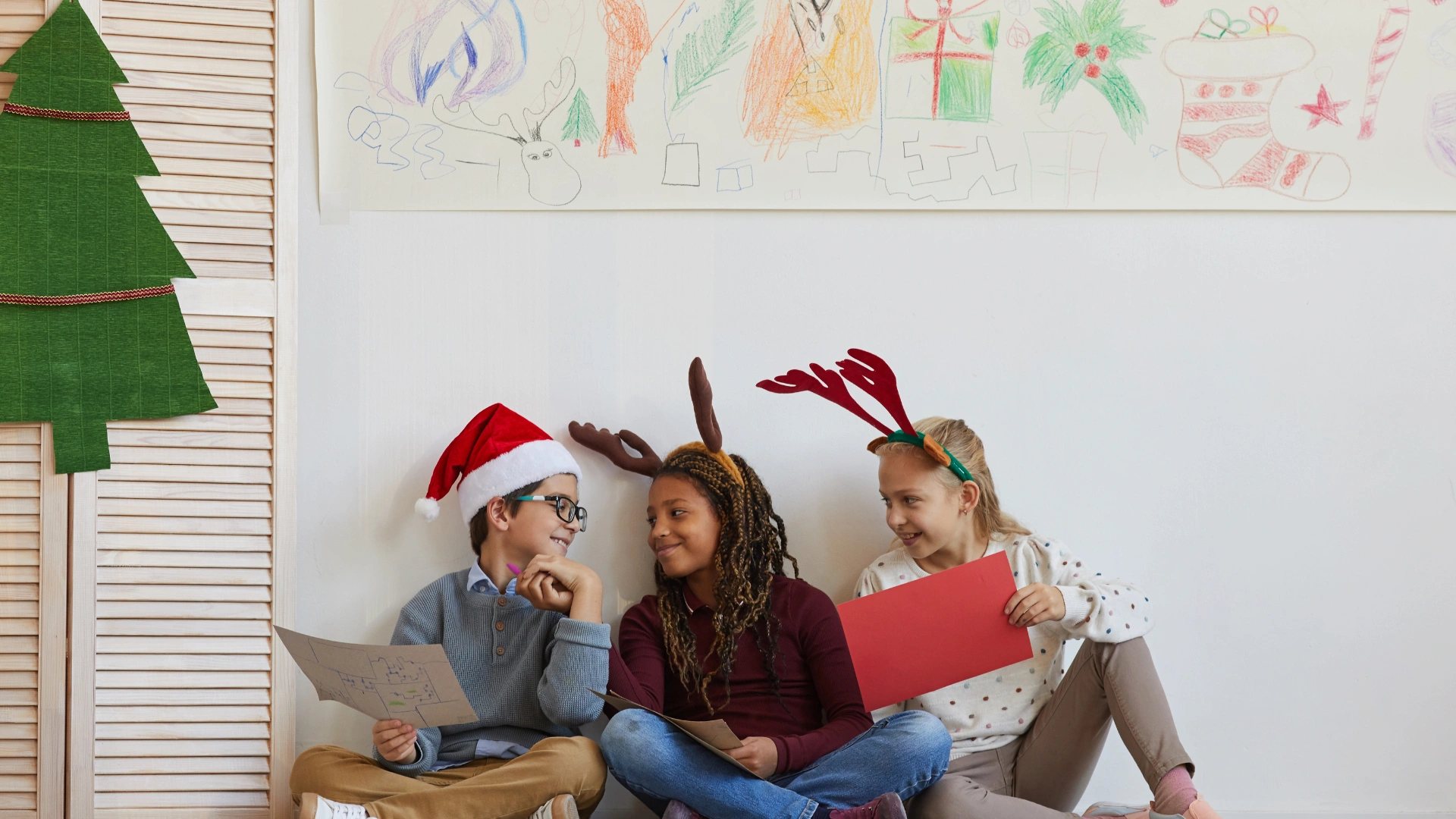 Three children sit on the floor wearing festive hats and antlers, smiling and holding papers. Behind them, a wall decorated with colorful holiday drawings sets a cheerful, playful mood—perfect for homeschooling through the holidays.