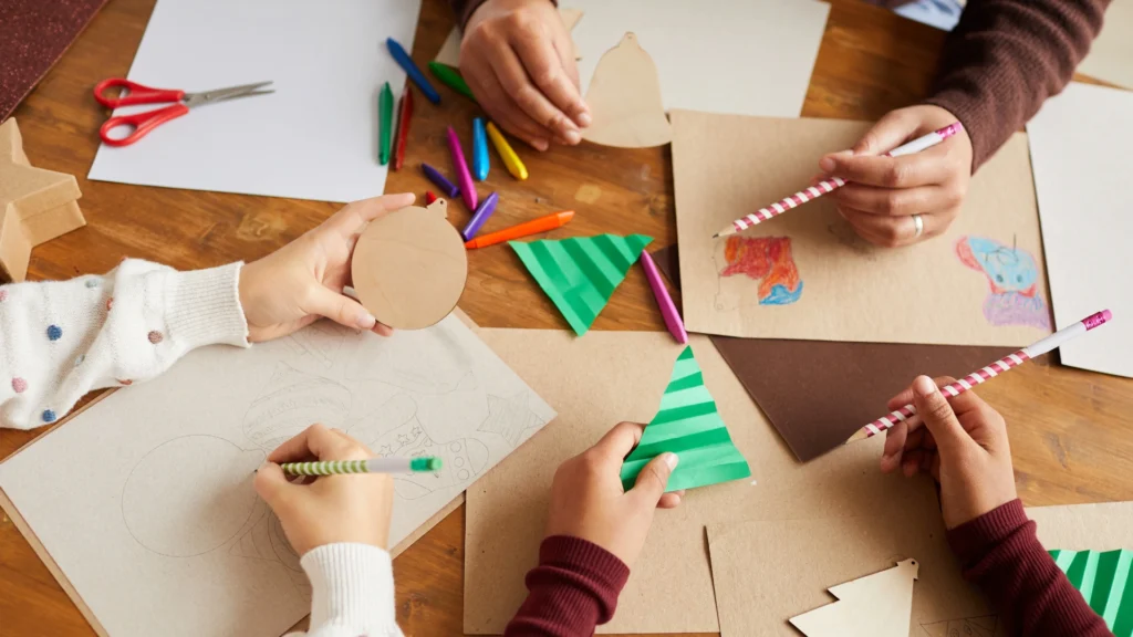 Four people work on holiday crafts at a table, drawing and coloring on brown paper shapes with colored pencils and crayons. Paper triangles, cutouts, and colorful supplies are scattered on the wooden surface.