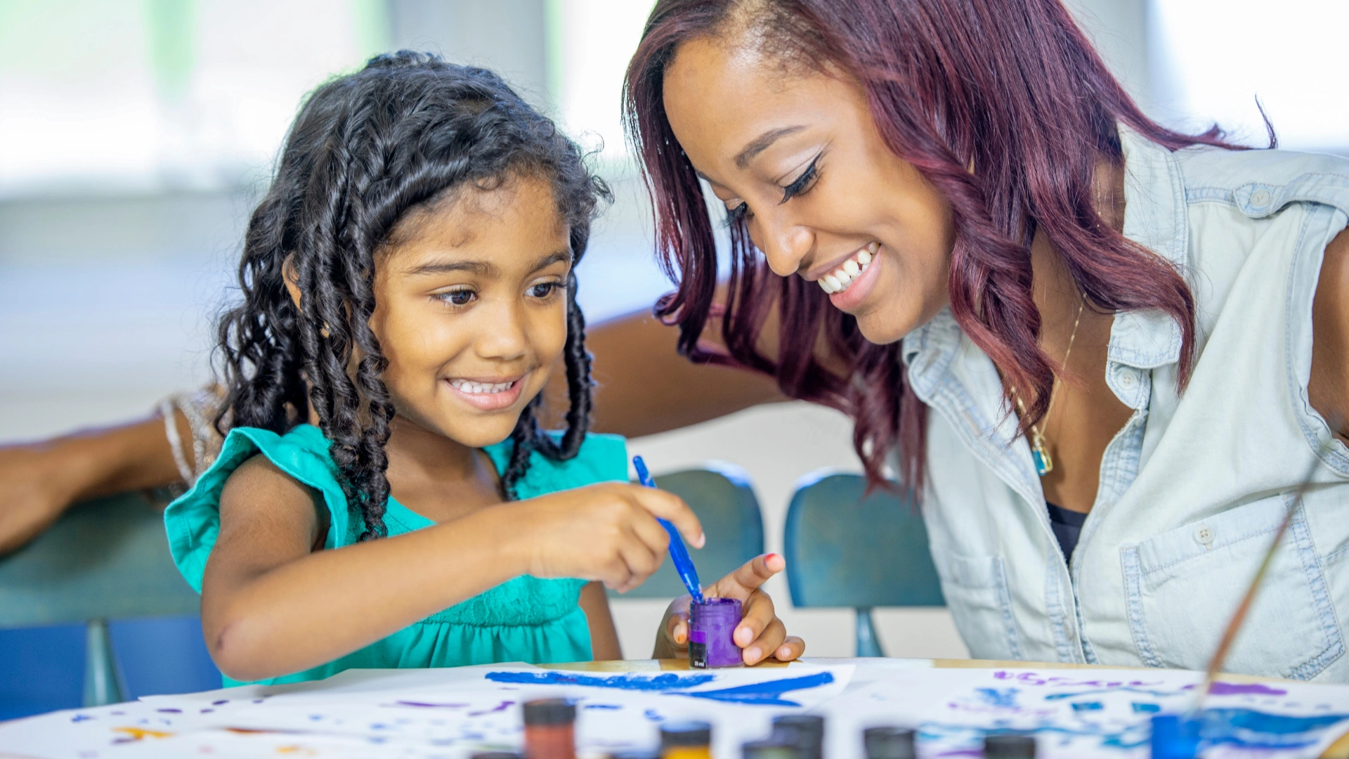 A young girl and a woman smile while painting together at a table, surrounded by colorful paint jars and paper, enjoying the freedom of a flexible homeschool schedule.