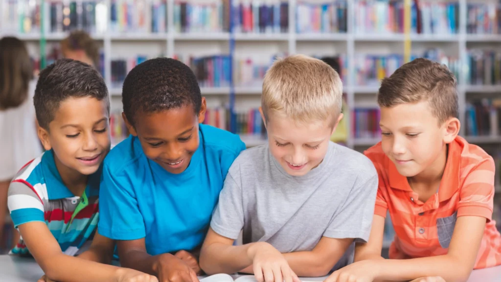 Four young boys sit side by side at a table in the Homeschool Resource Library, smiling and reading a book together, with bookshelves filled with books in the background.