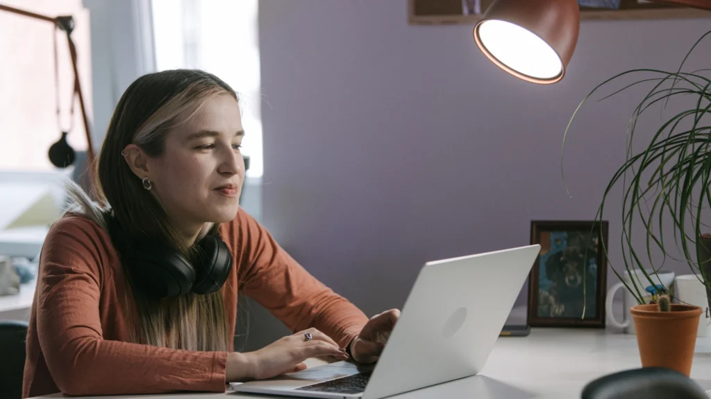 A person with long hair and headphones around their neck sits at a desk, using a laptop. As they appear focused on the screen, they are evaluating online homeschool programs. A lamp, potted plant, and framed photo complete the scene.