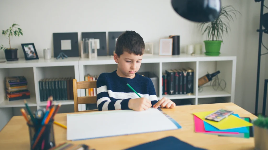 A young boy sits at a desk drawing on a large sheet of white paper with colored pencils. Books and plants line the shelves, creating a cozy study area ideal for spotting homeschool curriculum gaps and nurturing creativity.