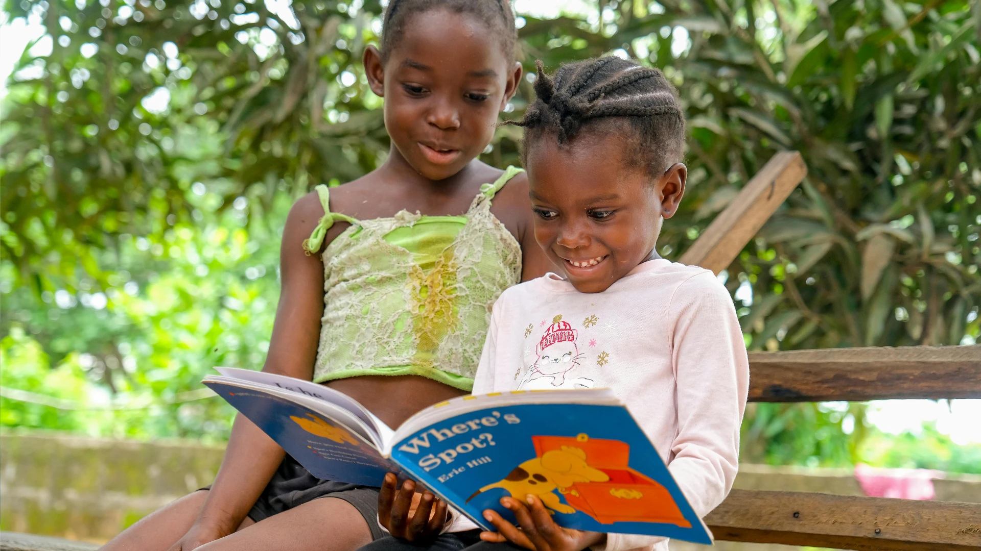 Two young girls sit outdoors on wooden steps, smiling and reading “Where’s Spot?” together—an engaging moment that supports homeschool reading fluency amid green trees and warm sunlight in the background.