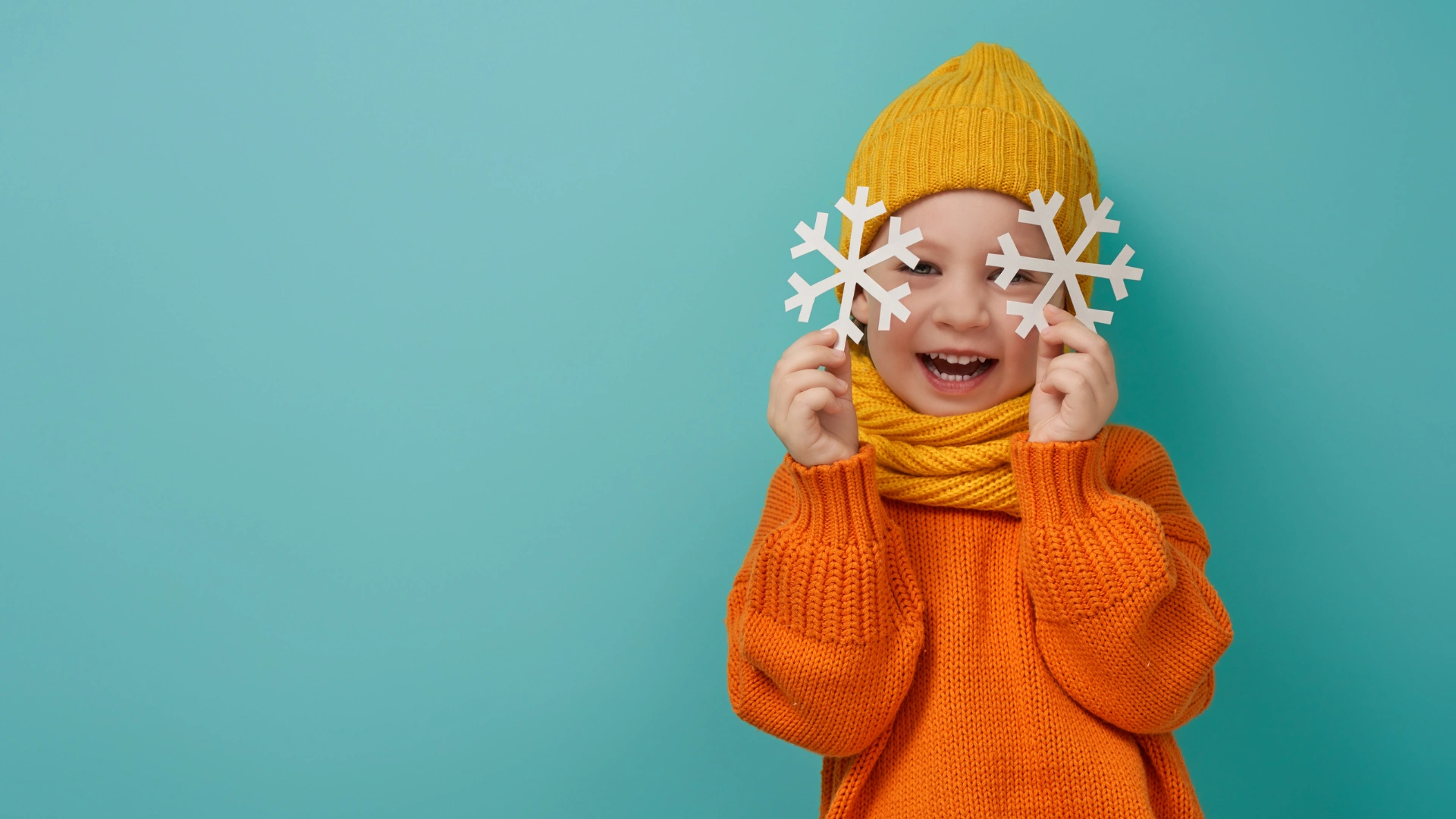 A smiling child in an orange sweater, yellow hat, and scarf holds two white paper snowflakes over their eyes, standing against a teal background—capturing the spirit of joyful learning and creativity.