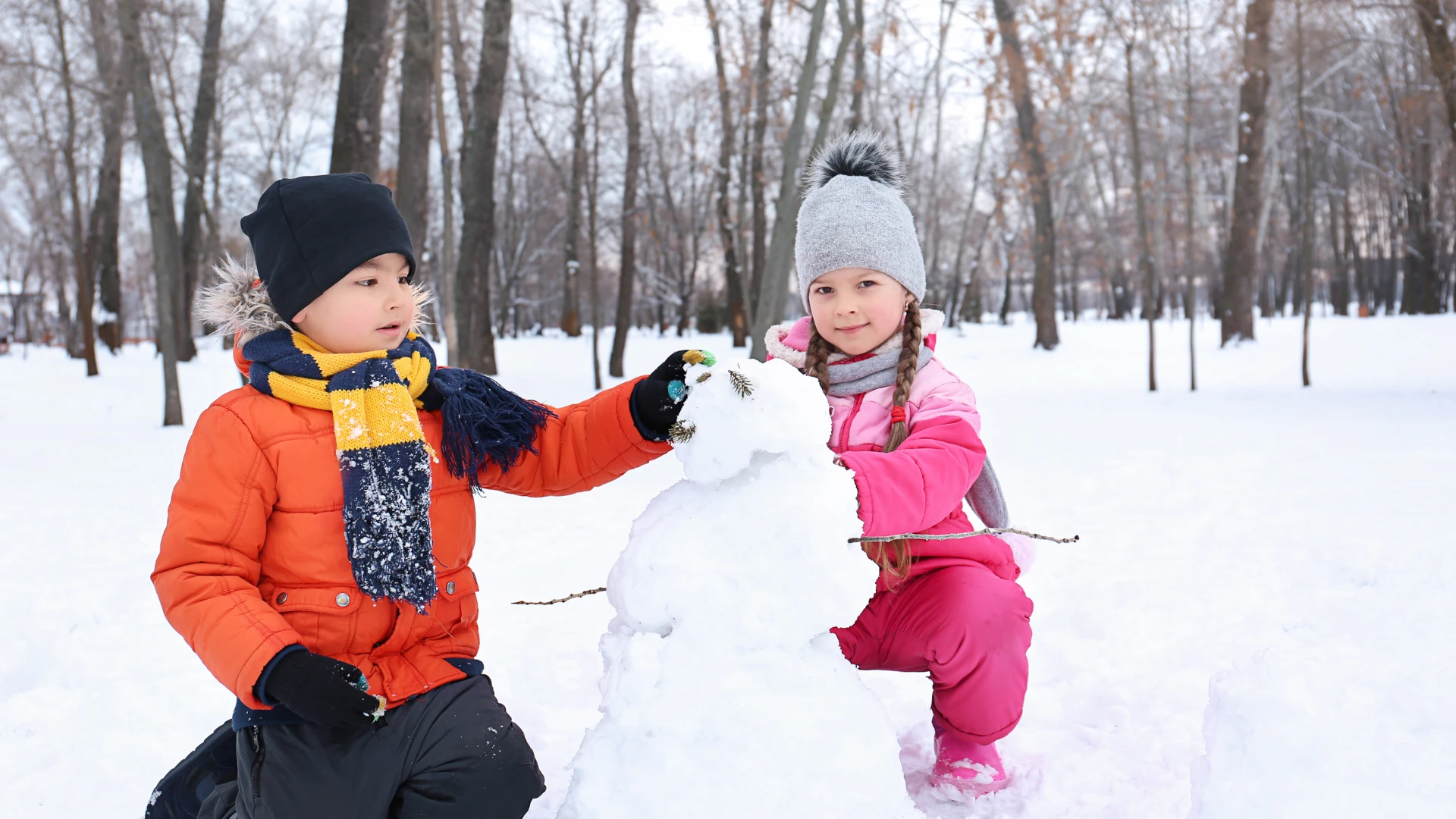 Two children dressed in winter clothes are building a snowman in a snowy park surrounded by bare trees, enjoying some hands-on Winter Homeschooling fun. The boy wears an orange jacket; the girl is in pink with a gray hat as they decorate the snowman's face.