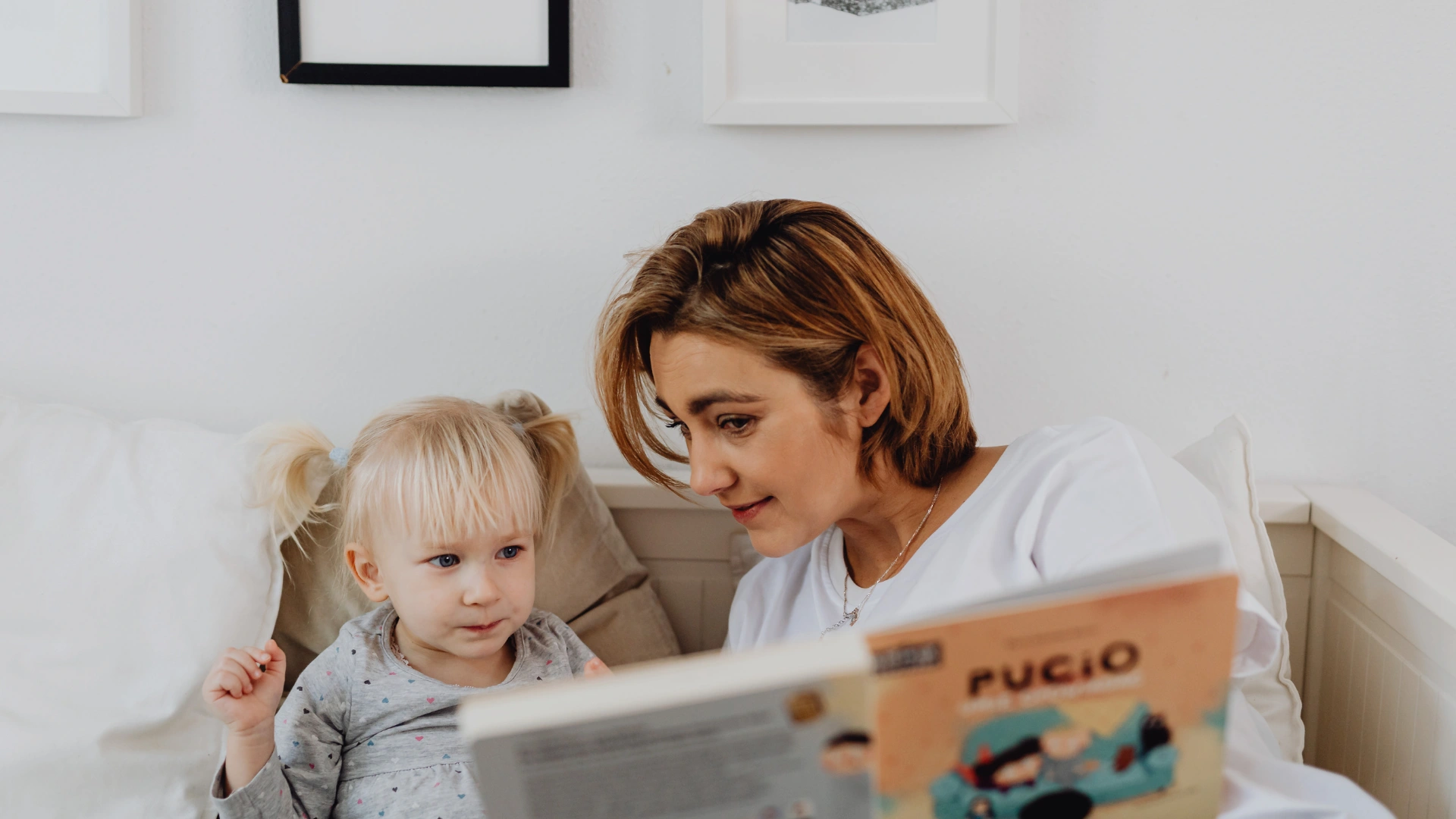 A woman with short blonde hair reads a picture book to a young girl with pigtails, both sitting on a couch in a cozy reading nook, surrounded by white walls and framed artwork in a well-lit room.