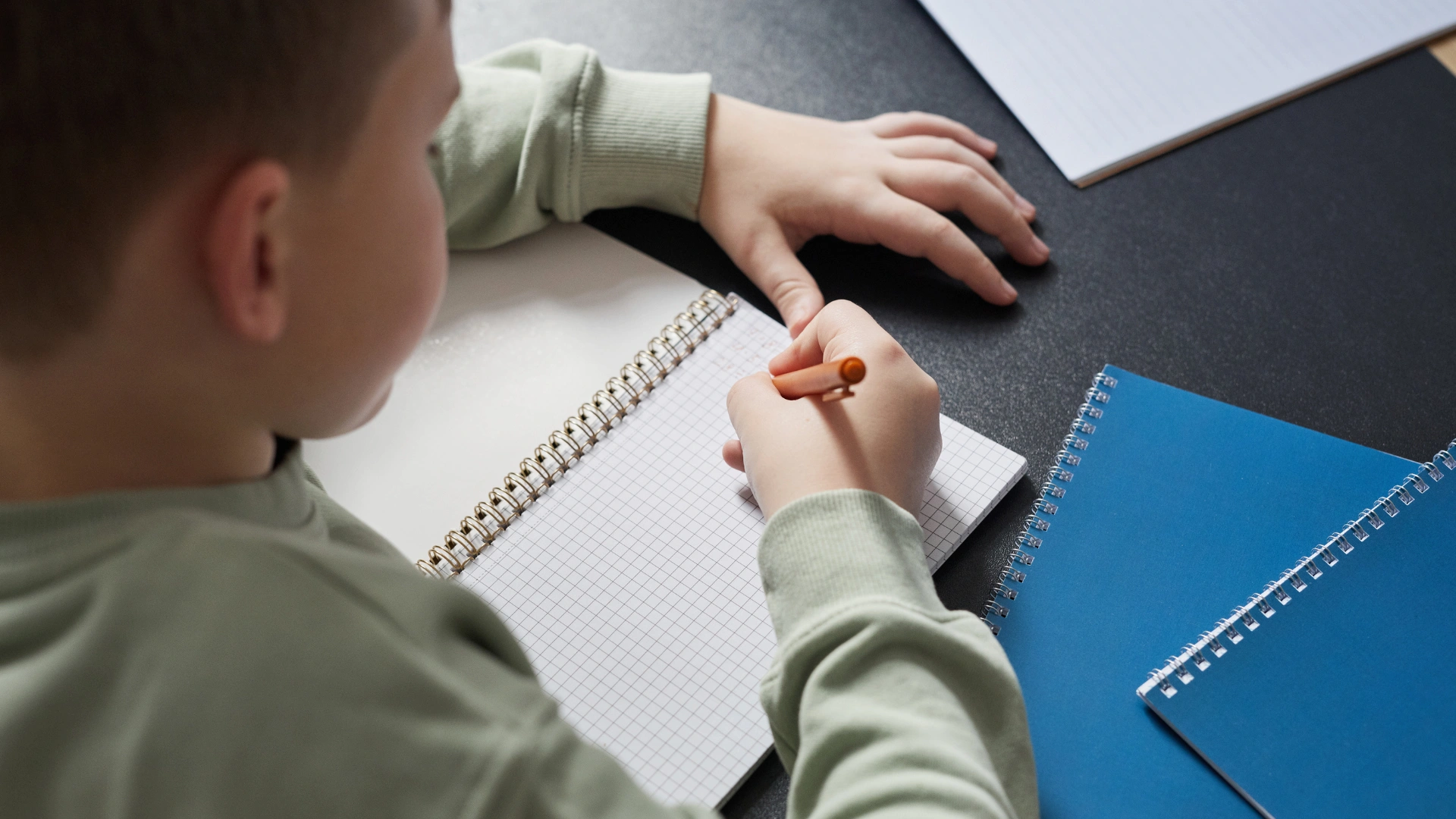 A child sits at a desk writing in a spiral notebook with a pencil—an encouraging scene for reluctant writers. Two other notebooks, one blue and one white, are nearby. The child wears a long-sleeved, light green shirt.
