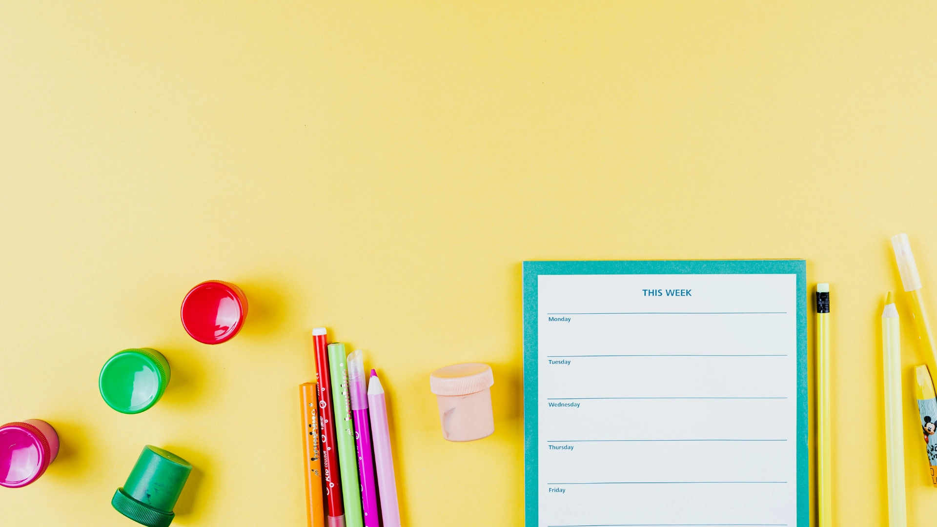 A weekly planner, colored pencils, pens, erasers, and a sharpener are arranged on a yellow background, creating a bright and organized workspace.