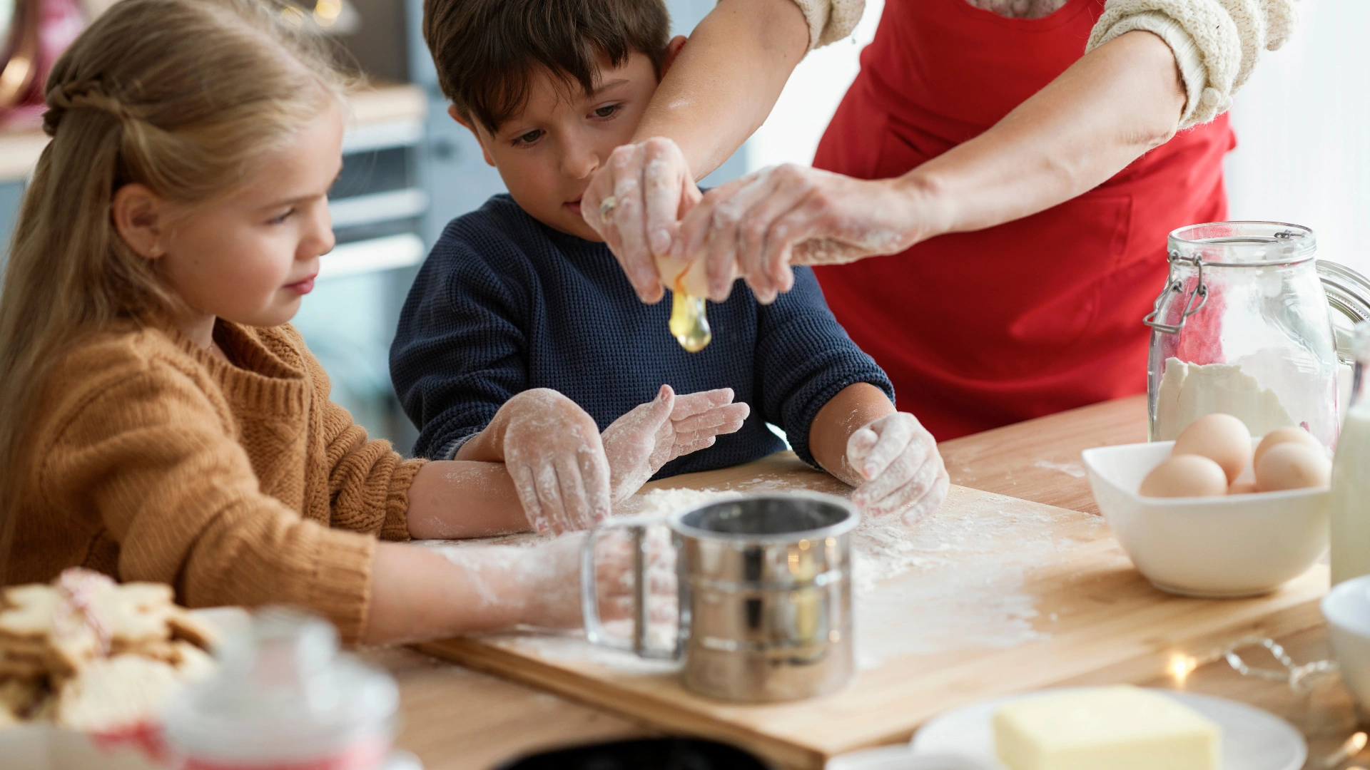 Two children watch as an adult cracks an egg over a floured surface in a kitchen. The kids have flour on their hands, and baking ingredients are on the table, suggesting they are baking together.