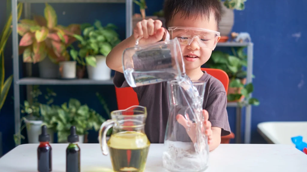 A young child in safety goggles pours water from a jug into a glass container on a table, creating a homeschool science lab scene. A pitcher of yellow liquid, dropper bottles, and green plants are visible in the background.