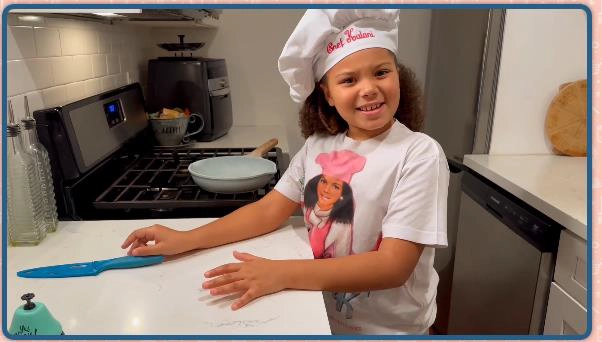 A young child wearing a chef hat and a t-shirt with a woman’s face on it stands and smiles at a kitchen counter with a blue-handled knife and a frying pan in the background.