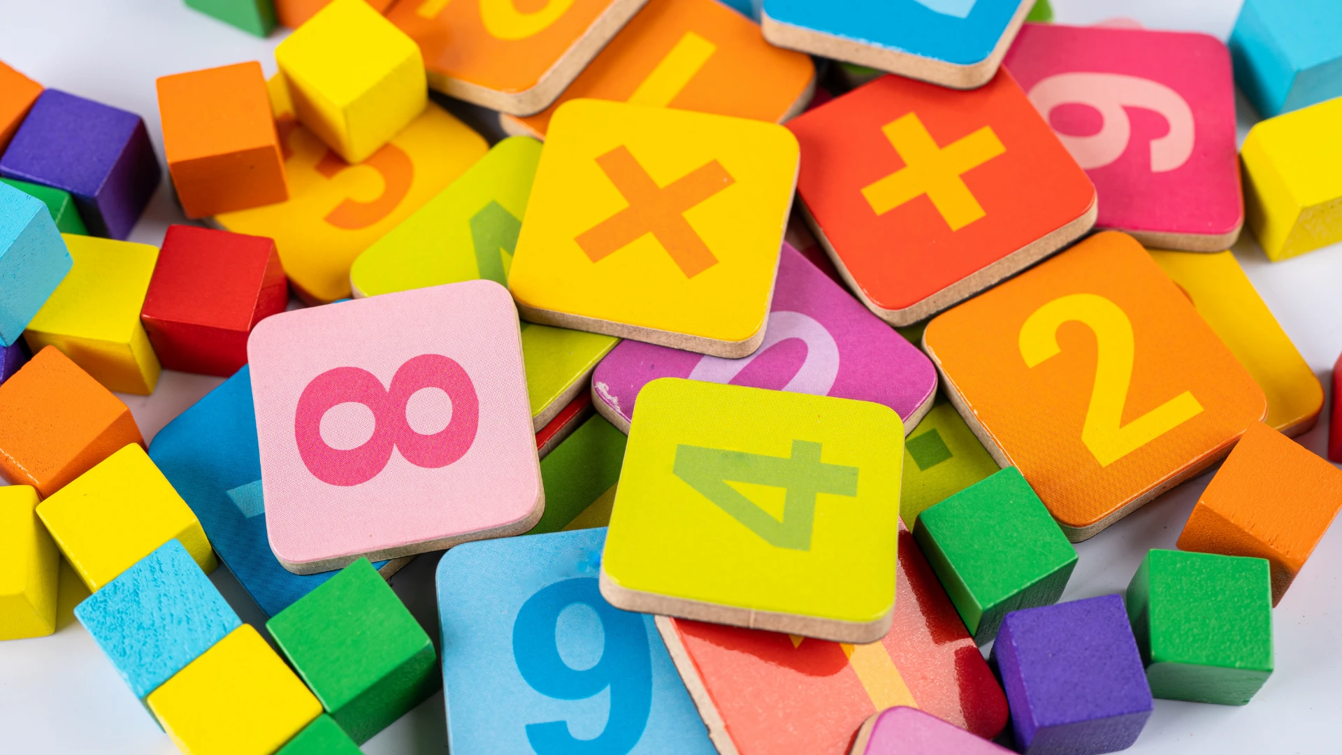 A colorful assortment of wooden blocks featuring numbers, plus signs, and squares, along with small multicolored cubes, ideal for real world math activities, scattered on a flat surface.
