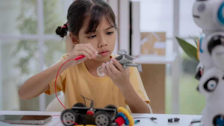 A young girl in a yellow shirt works on a small robot with wires and tools at a table, focusing intently while applying simple engineering concepts to assemble or repair the device in a bright room.