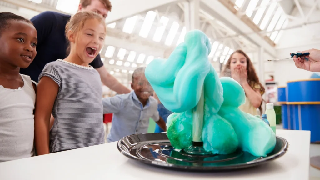 A group of excited children and an adult watch a science experiment, where foaming green-blue liquid erupts from a bottle on a tray in a bright, spacious room.