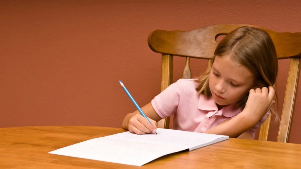 A young girl with light brown hair sits at a wooden table, wearing a pink polo shirt, and writes with a pencil on paper, resting her head on one hand and concentrating on her work.