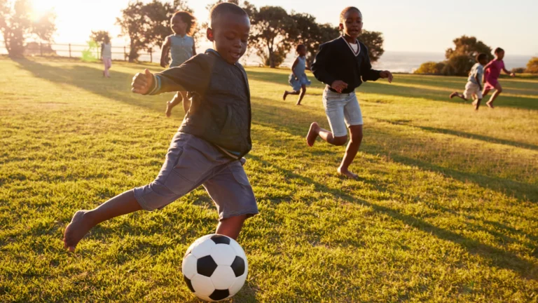 Four children play soccer on a grassy field at sunset, making the most of extracurricular ideas for homeschoolers. One boy in front is about to kick the ball while others run behind him, enjoying the game. Trees and the sun are visible in the background.