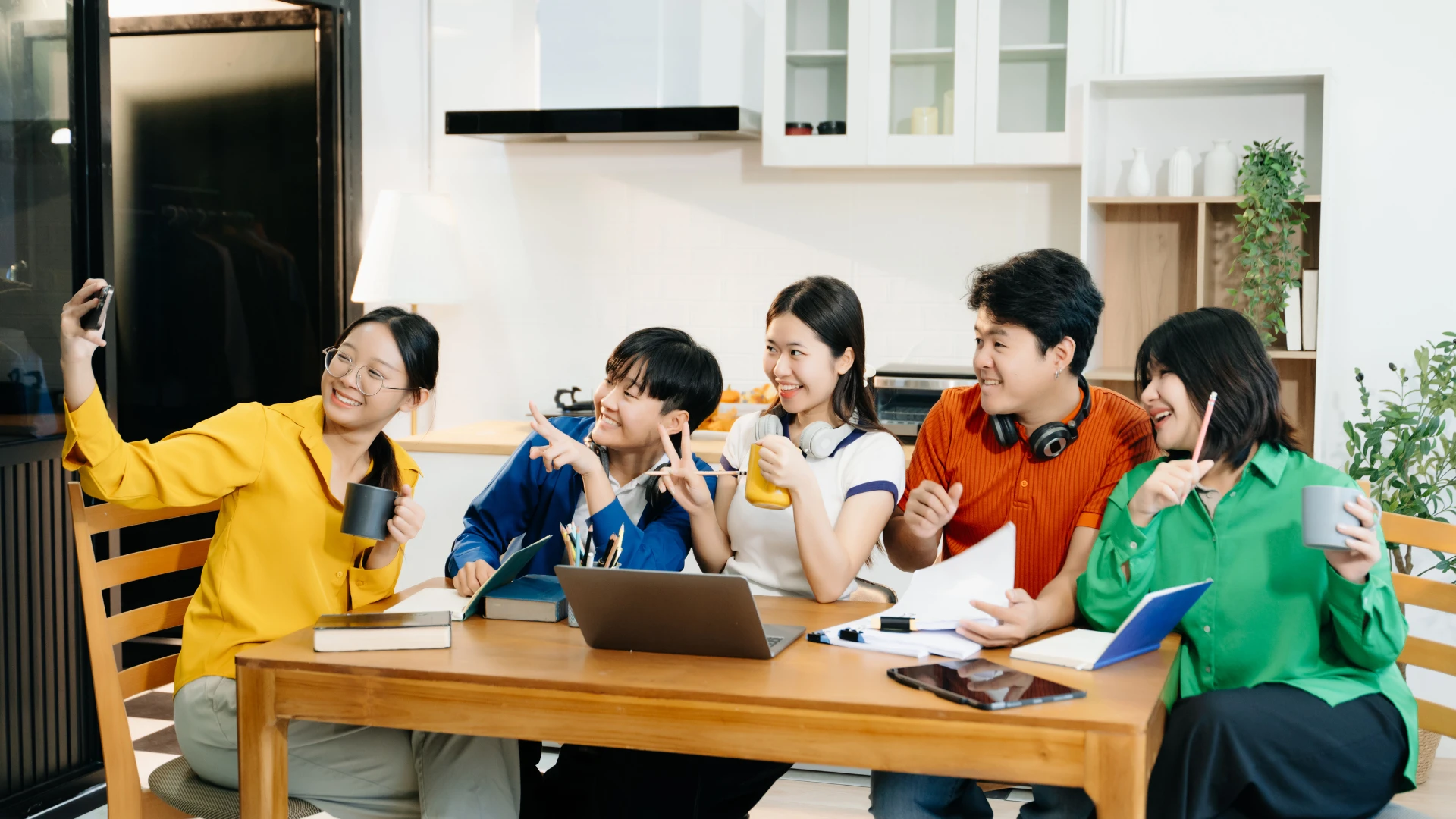 Five young adults sit around a table, smiling and posing for a selfie. Surrounded by books, a laptop, and mugs, they appear cheerful—capturing the collaborative spirit you’ll find when you Start Your Own Homeschool Co-op.