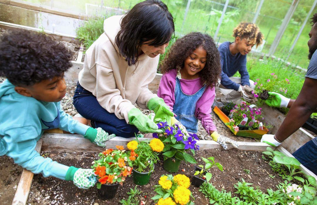 A group of children and an adult garden together in a raised bed, planting colorful flowers and smiling. Everyone wears gardening gloves and enjoys the outdoor activity in a lush, green setting.