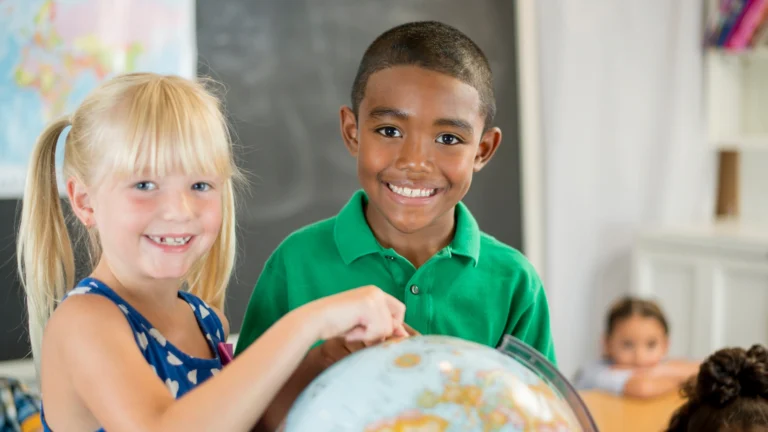 Two smiling children stand next to each other in a classroom, pointing at a globe—a perfect introduction to teaching geography at home. A girl with blonde hair and a boy in a green shirt look at the camera, ready to explore new places. A blurred blackboard and classmates are visible in the background.