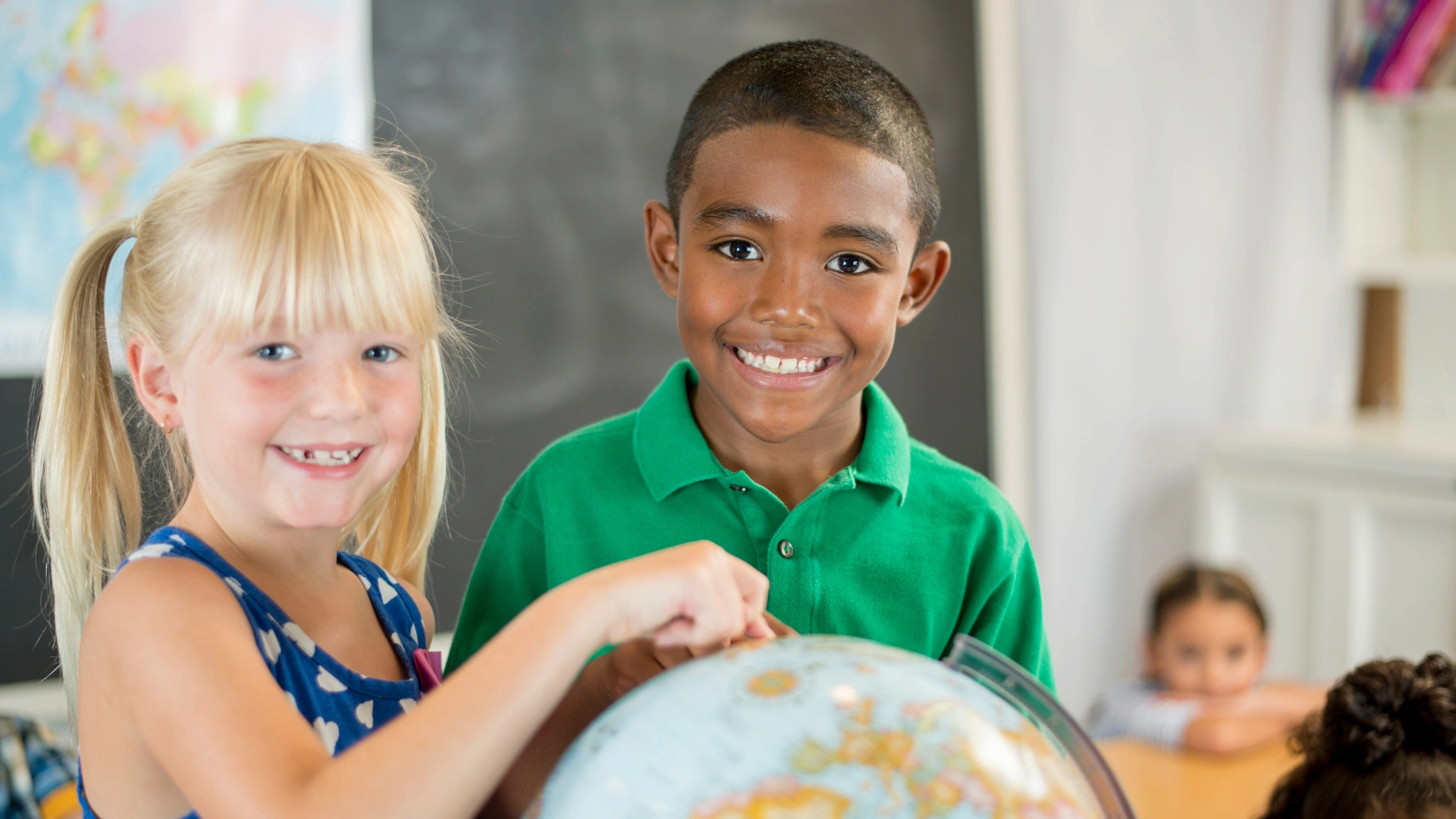 Two smiling children, one girl with blonde hair and one boy with short dark hair, stand together in a classroom, pointing at a globe—an inspiring scene for Teaching Geography at Home. A blackboard and other students are visible in the blurred background.