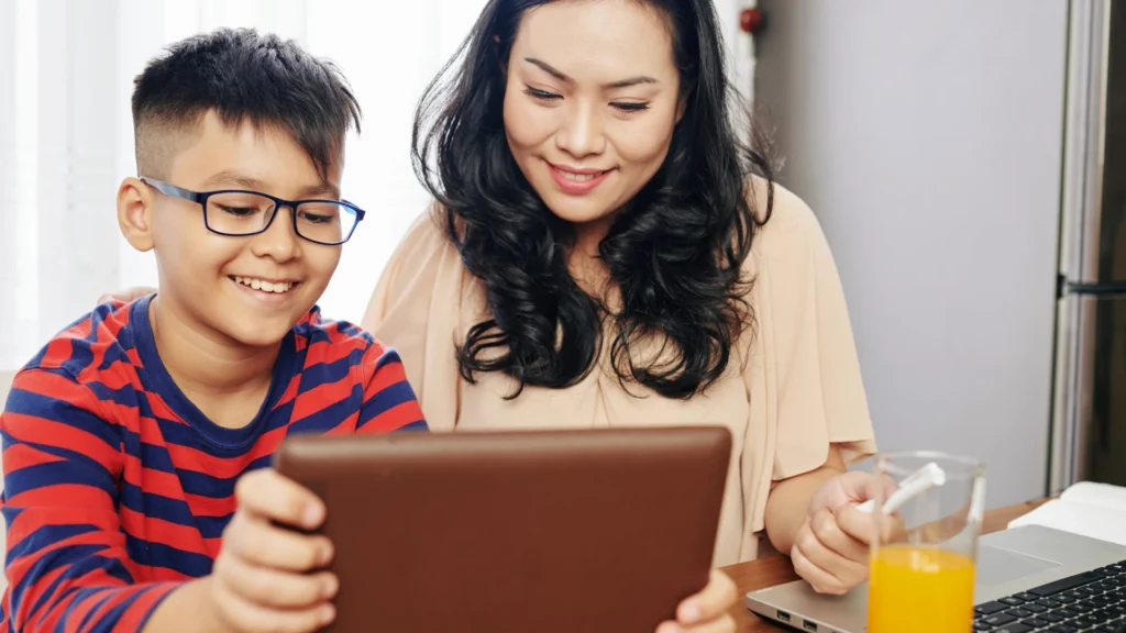 A smiling woman and a boy with glasses are sitting at a table, looking at a tablet together. The boy wears a red and blue striped shirt. There is a laptop and a glass of orange juice on the table.