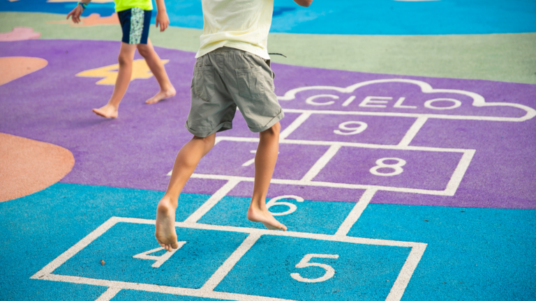 Two barefoot children play hopscotch on a colorful outdoor court, discovering how to keep kids learning in summer through playful movement and counting. The hopscotch grid is labeled with numbers and the word CIELO inside a cloud at the top, blending fun with learning under the open sky.