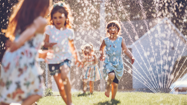 Four young children run and play through a backyard sprinkler on a sunny day, splashing water as they laugh and enjoy the outdoors. The water droplets sparkle in the sunlight, offering both joyful play and a perfect break to support summer learning loss prevention.
