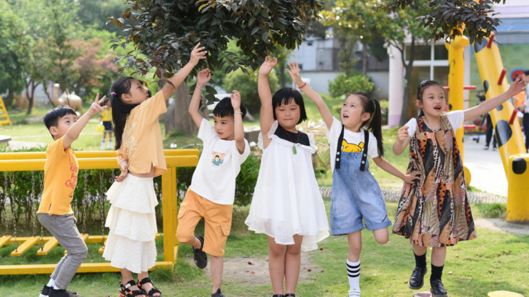 Six children stand outdoors reaching up toward tree branches, smiling and playing together. The scene is bright and sunny, with green grass and playground equipment visible in the background—a joyful reminder that while questions like “how much should kids study in summer” may arise, playtime is just as important for their growth and happiness.