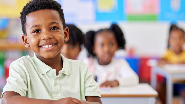 Smiling young boy sits at a classroom desk, looking at the camera, as if inviting the question, “is my child on track academically?” Other children are visible behind him, slightly out of focus, in a colorful classroom setting.