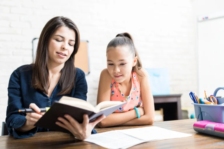 A woman is sitting at a table reading a book and showing it to a young girl, who is listening attentively. Papers and school supplies are on the table in front of them as they discuss How to Use Online Reading Programs for Summer Joy.