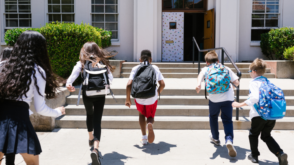 Five children with backpacks run up the steps toward the entrance of a school building on a sunny day.
