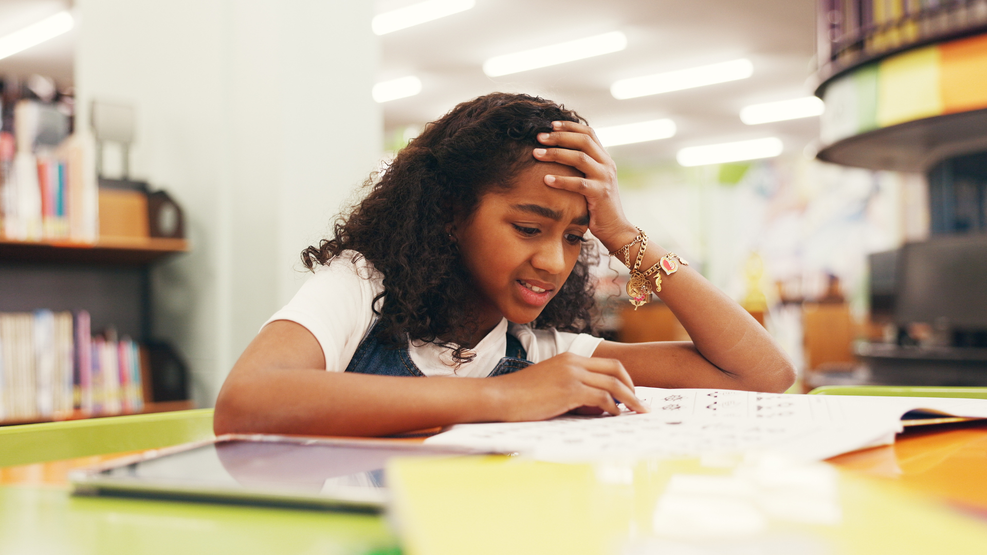 A young girl sits at a table in a library, looking stressed as she studies a worksheet, resting her forehead on one hand and frowning in concentration—perhaps feeling overwhelmed by the pressures of keeping up with her school routine for kids.