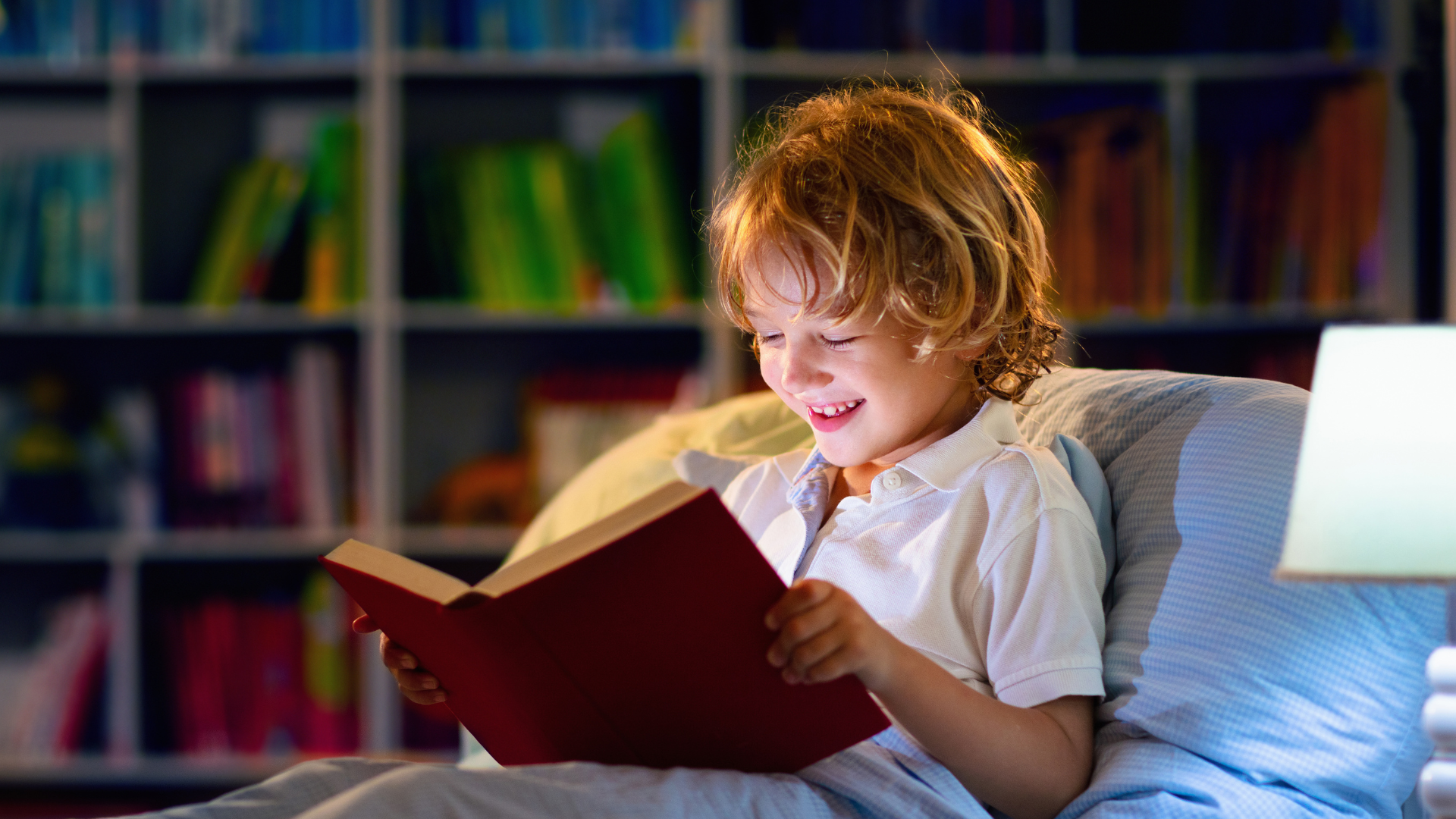 A young child with curly hair sits in bed, smiling and reading a red book. A lamp glows nearby, and a bookshelf filled with colorful books is in the background.