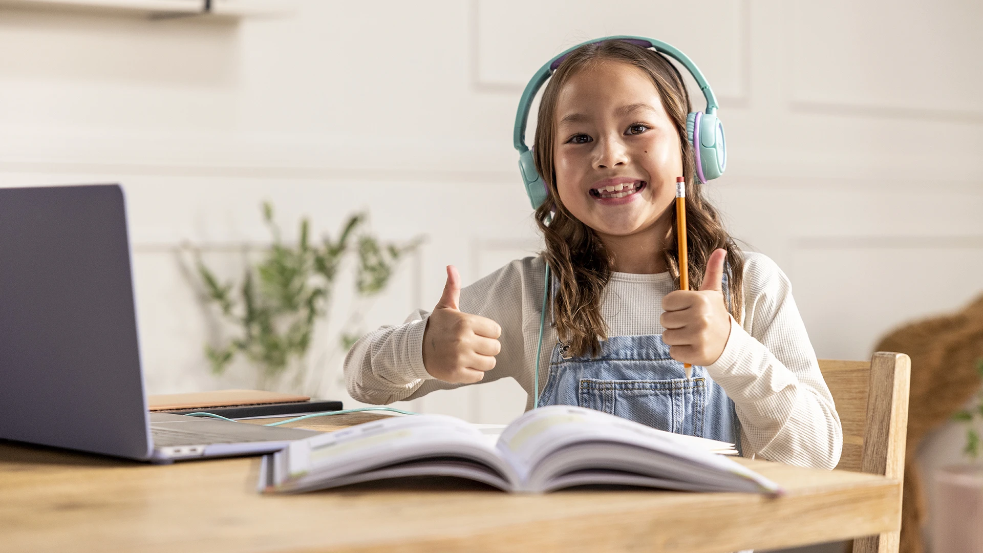 A smiling child wearing headphones gives two thumbs up while sitting at a table with an open book and a laptop, suggesting online learning or homework—perhaps enjoying a fun session with an online math coach.